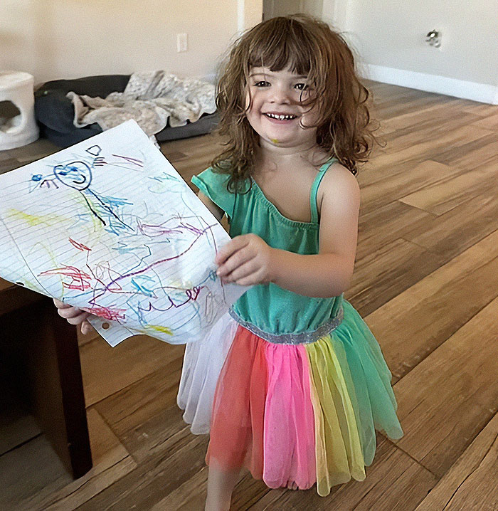 Toddler in colorful dress holding a drawing indoors, representing a 2-year-old daughter in hot car tragedy story.