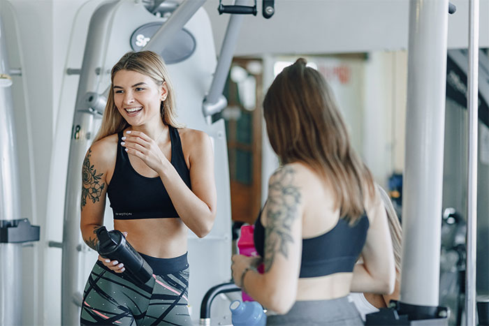 Two young women with tattoos in athletic wear smiling and talking at the gym during a workout session.
