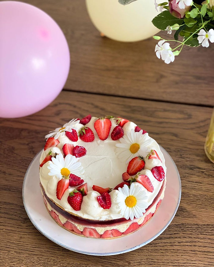Strawberry and cream cake decorated with daisies on a wooden table with balloons and flowers nearby, teen cake fail theme.