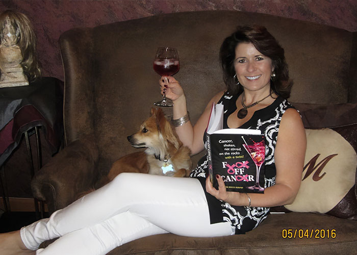 Woman holding book and glass of wine, sitting on couch with dog, reflecting on funny obituary she wrote herself.