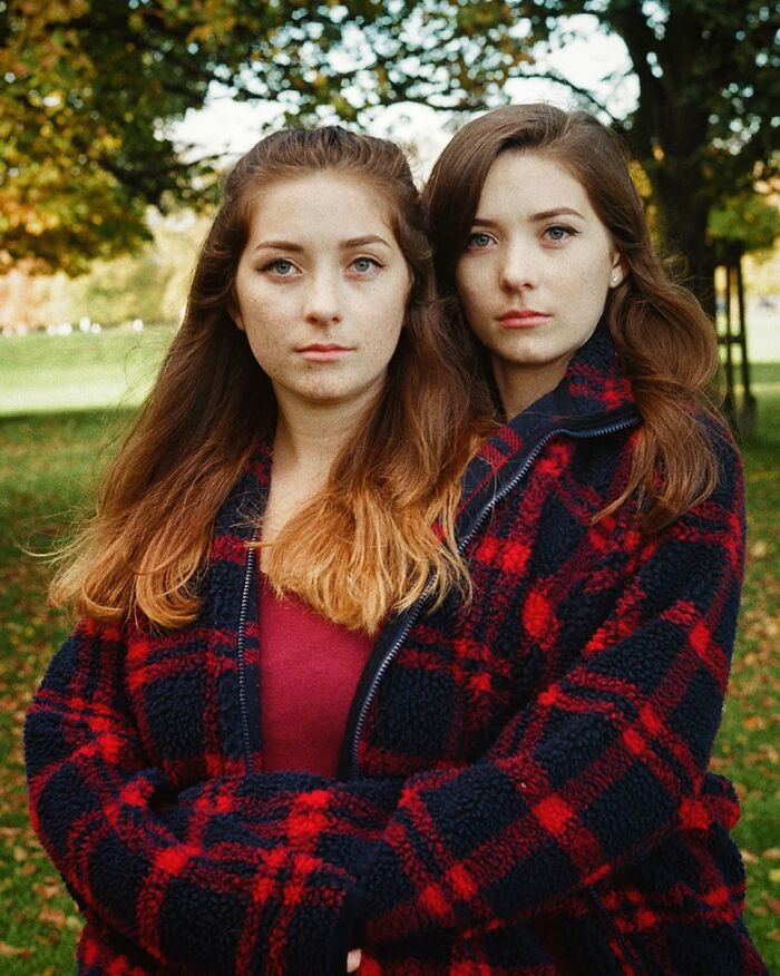 Twin sisters wearing matching red and black plaid jackets posing closely in a park, captured by a London photographer.