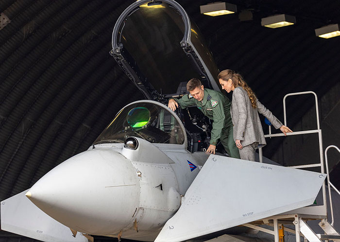Servicemen inspecting jet cockpit with Kate Middleton standing nearby inside military aircraft hangar.