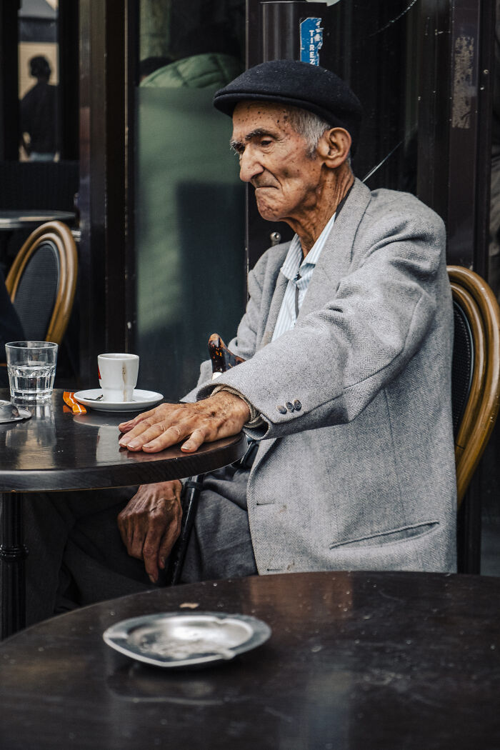 Elderly man in a hat sitting at a café table, captured in stunning street photography of life’s unexpected moments.