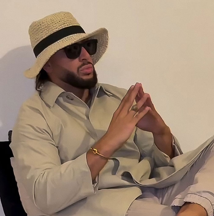 Man wearing sunglasses and a straw hat sitting with hands clasped, linked to influencer fake syringe attack pranks case.