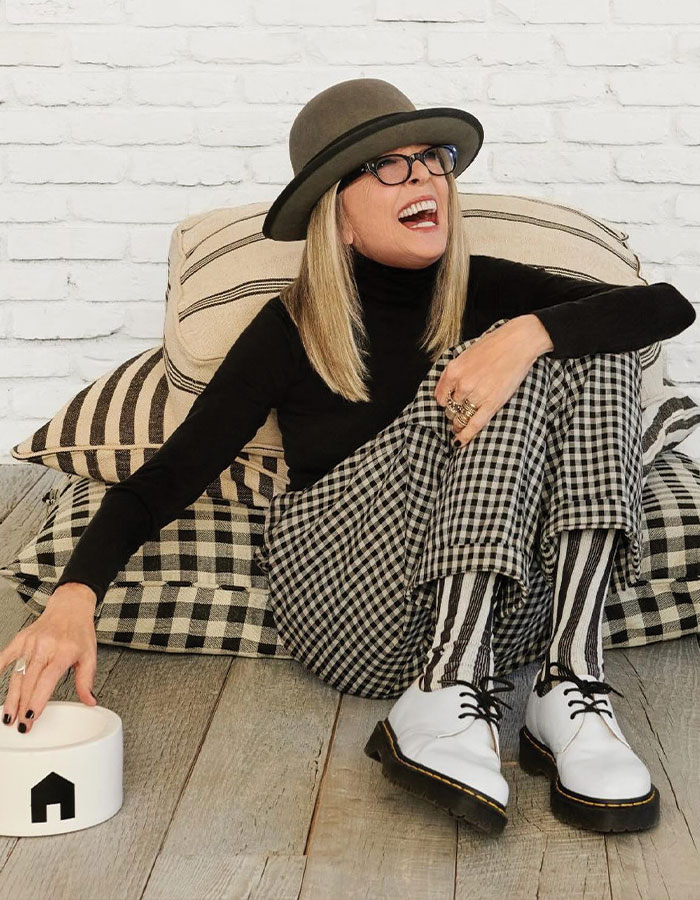 Diane Keaton sitting on pillows in patterned pants and white shoes, reflecting on health decline before selling $28M home.