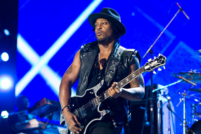 R&B legend D'Angelo performing on stage wearing a black hat and leather vest with electric guitar.