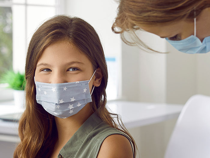 12-year-old girl wearing a star-patterned mask sitting in a medical office with a masked adult attending to her.