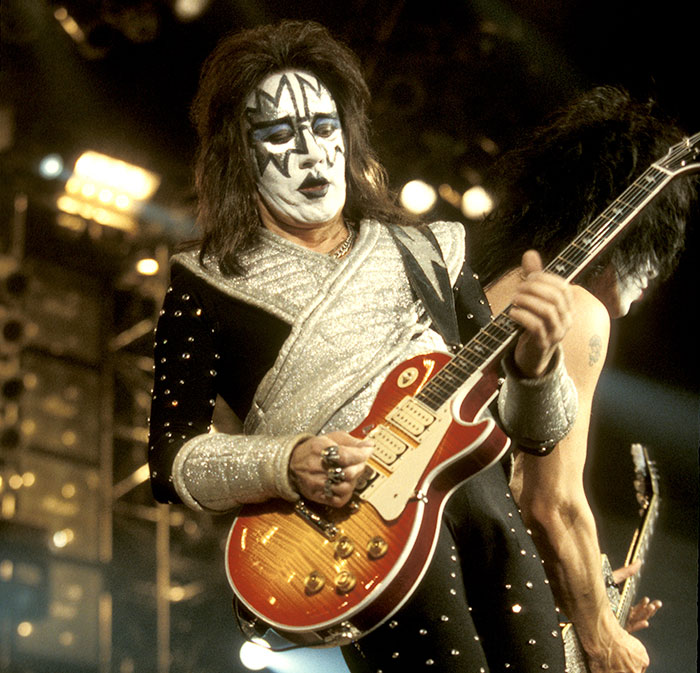 Gene Simmons performing with Kiss on stage, wearing signature makeup and playing a sunburst electric guitar.