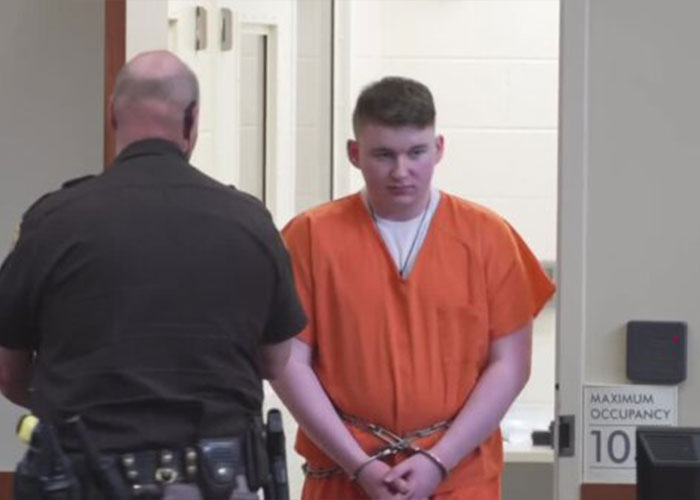 Teen dad in orange prison jumpsuit handcuffed, facing officer in black uniform inside a detention facility hallway.