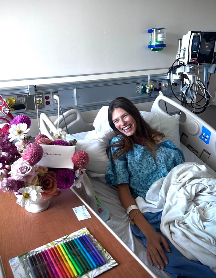 Ex&ndash;Victoria&rsquo;s Secret model smiling in hospital bed, battling cancer with flowers and markers on nearby table.