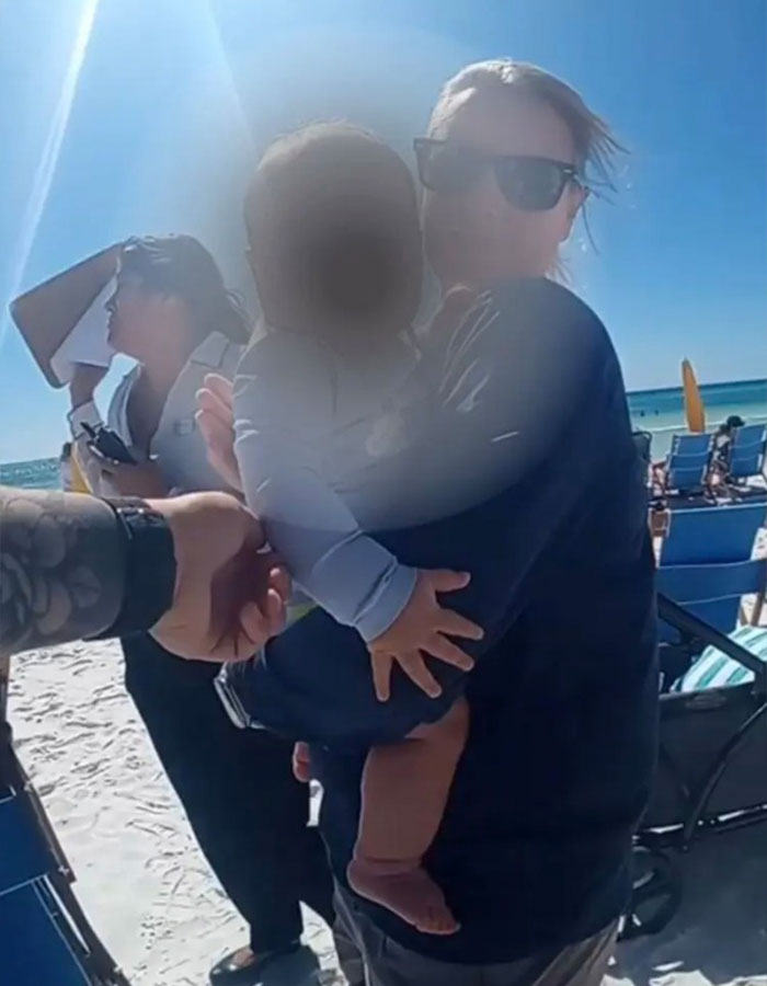 Woman holding 6-month-old baby on beach near people in chairs on sunny day, highlighting major twist case.