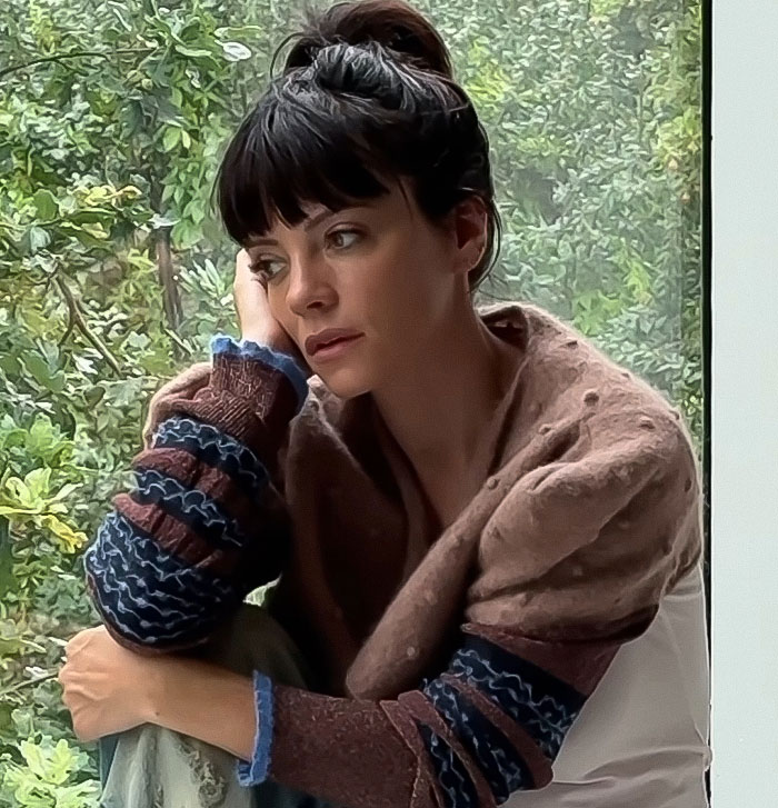 Woman with dark hair in a bun sitting by a window, appearing thoughtful amid discussions about David Harbour cheating controversy.