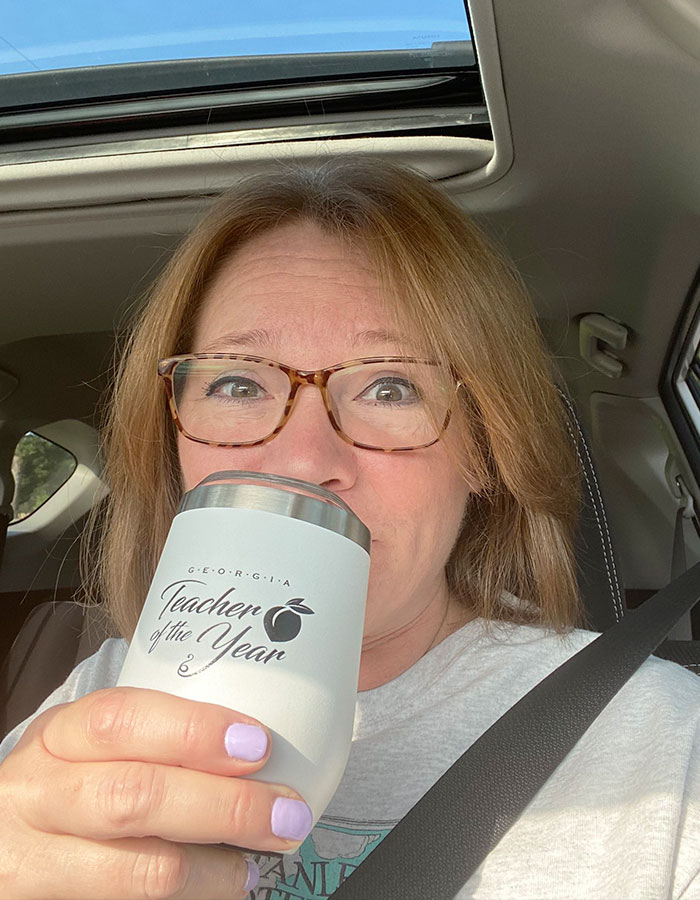 Woman with glasses holding a Teacher of the Year cup inside a car, related to Teacher Of The Year finalist news.