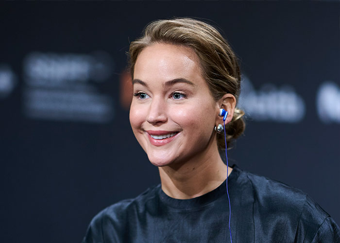 Jennifer Lawrence smiling during a public event wearing a black top and blue in-ear headphones discussing plastic surgery plans.