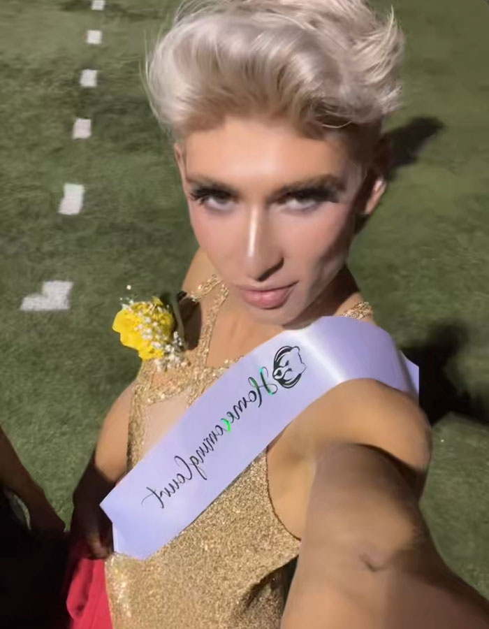 Young man wearing a gold dress and homecoming queen sash taking a selfie on a sports field after his viral win.