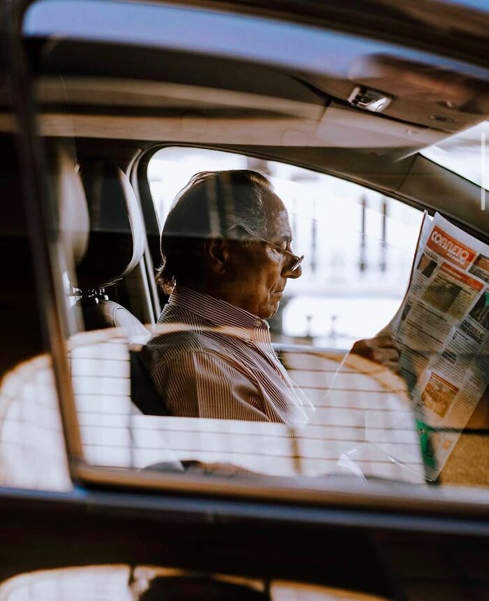 Elderly man reading a newspaper inside a car, capturing the beauty and wisdom of old age in a street photo.
