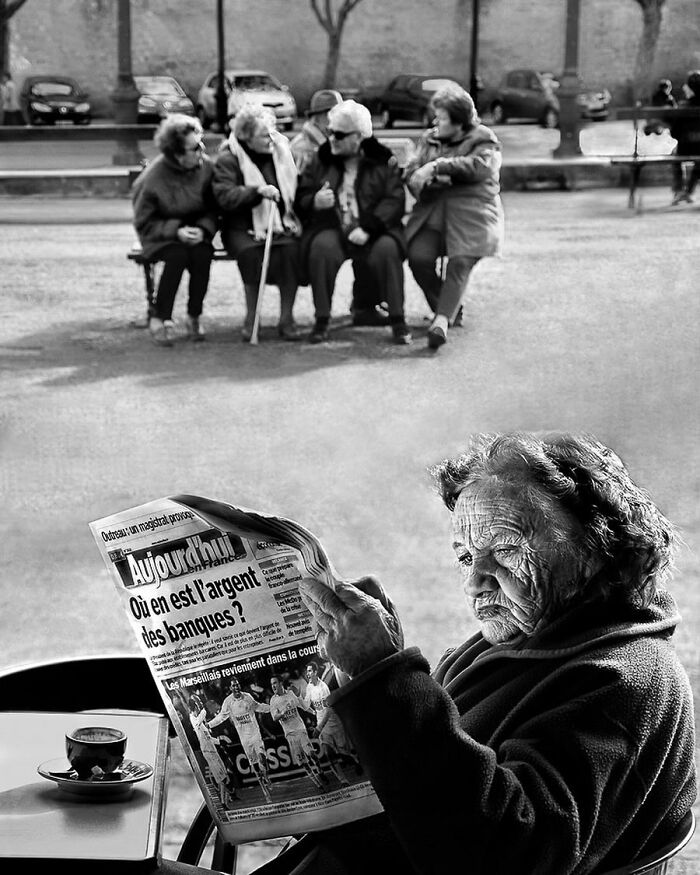 Elderly woman reading a newspaper at a cafe with a group of older adults socializing on a bench in the background.