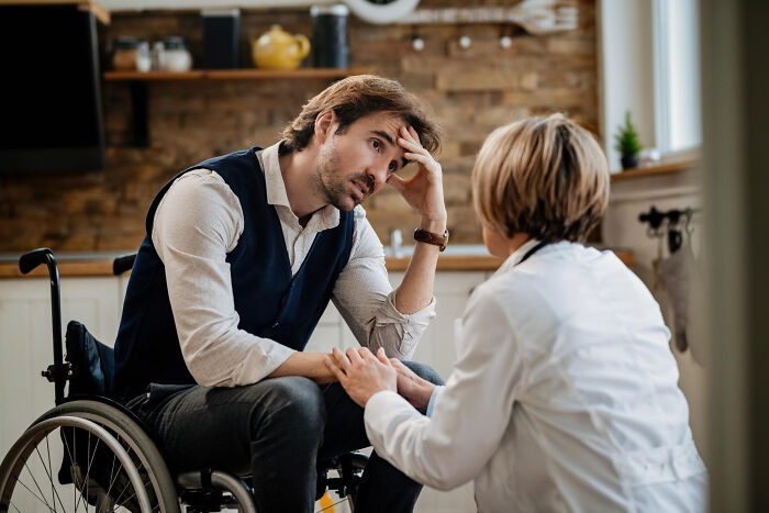 Man in wheelchair looking distressed while a healthcare professional comforts him in a home setting, showing disability support.
