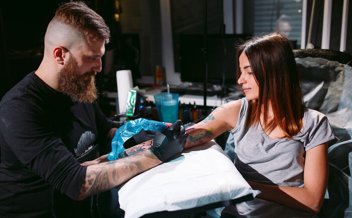 Tattoo artist with gloves working on a woman's colorful arm tattoo in a professional tattoo studio setting.