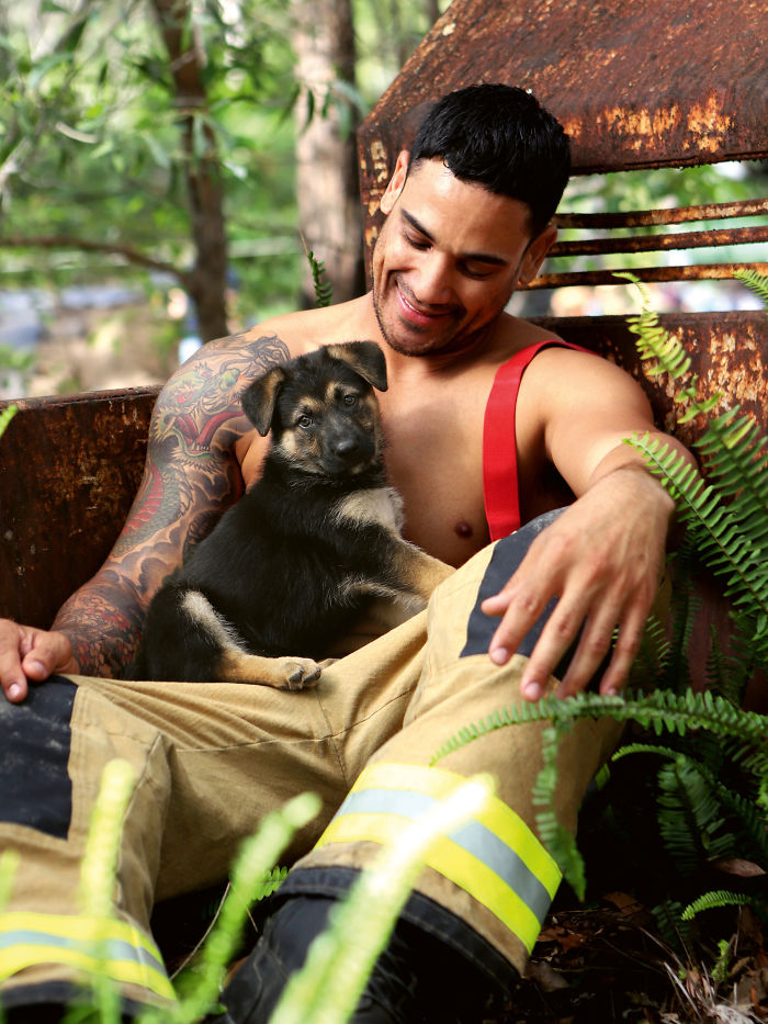Smiling firefighter in pants and suspenders holding an adorable dog, surrounded by greenery and rustic metal background.