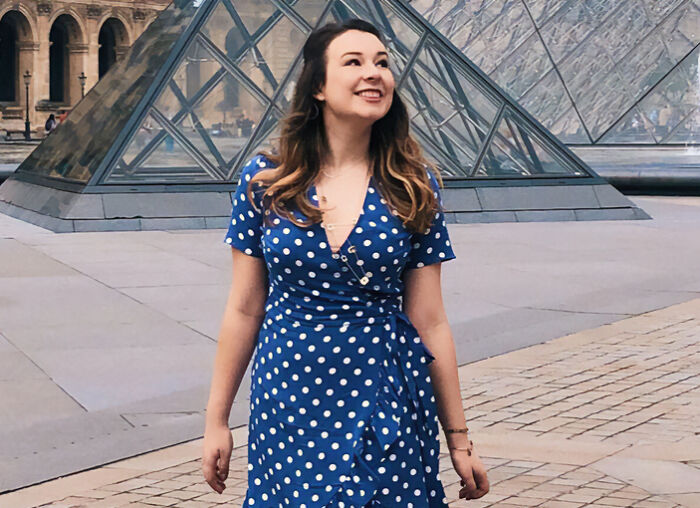 Young woman in a blue polka dot dress smiling near glass pyramid, related to Disney superfan life update and news.