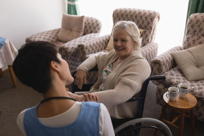 Elderly woman in a wheelchair smiling and talking with a caregiver in a cozy living room, illustrating dementia patient interaction.