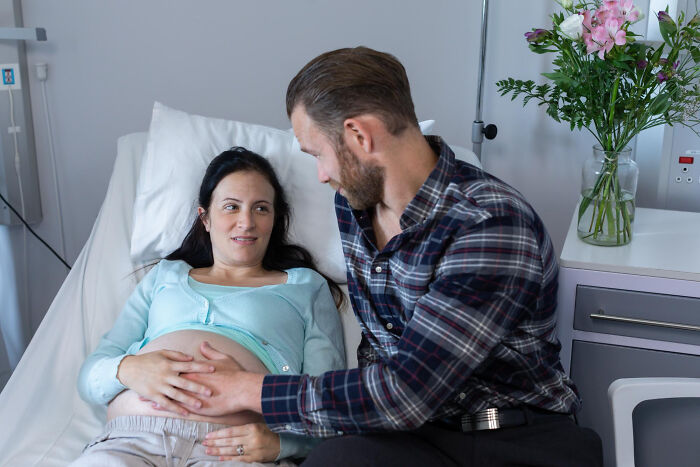 Expectant dad touching pregnant wife's belly in hospital room, showing lovable clueless dad moments during pregnancy.