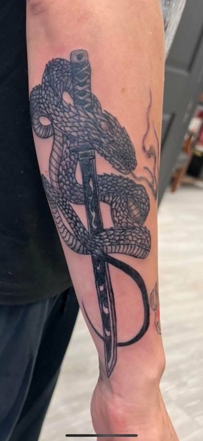 Black ink tattoo of a coiled snake wrapped around a katana blade on a forearm, showcasing intricate scales and design.