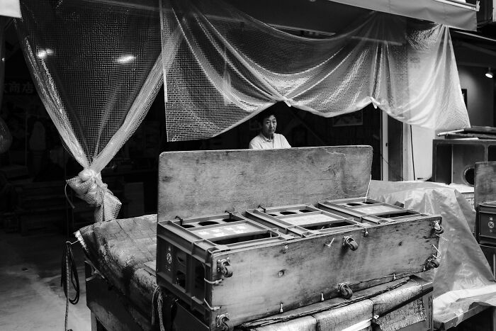 Black and white street photography capturing a man sitting behind wooden crates in an urban market setting.