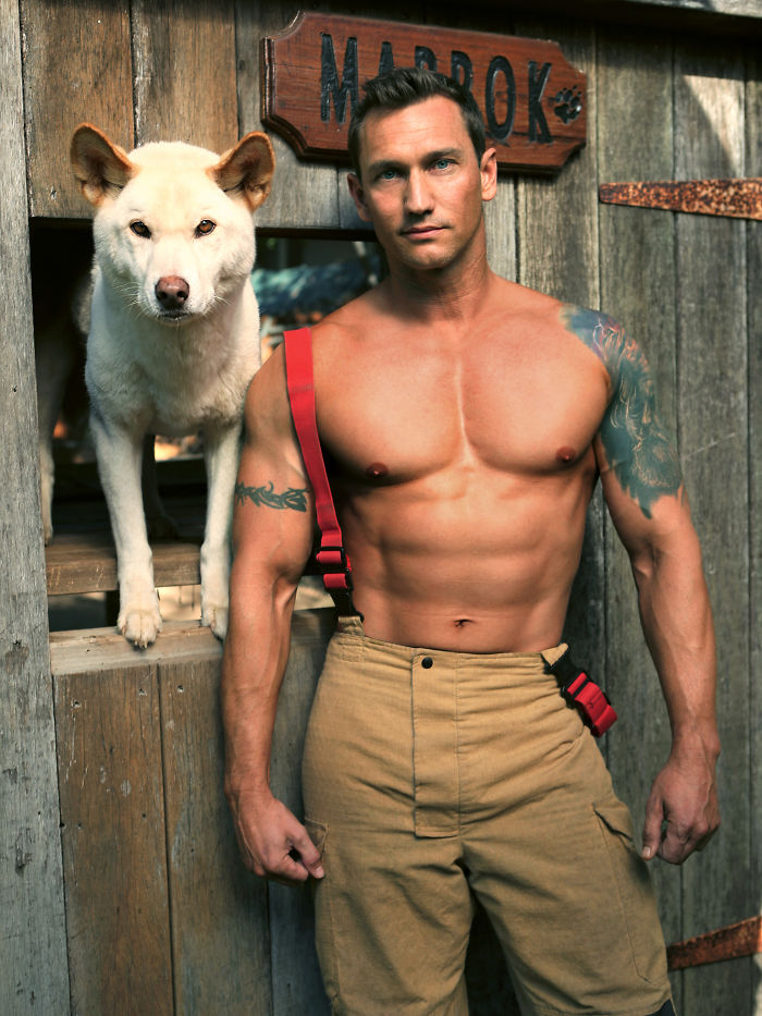Shirtless heroic firefighter with tattoos standing next to an adorable dog in front of a rustic wooden door.