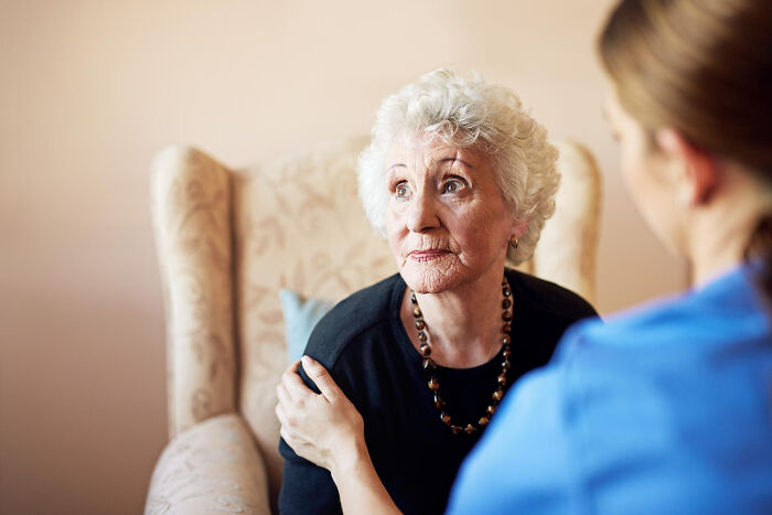 Elderly woman with dementia sitting in a chair, looking thoughtful while a caregiver offers support and comfort.
