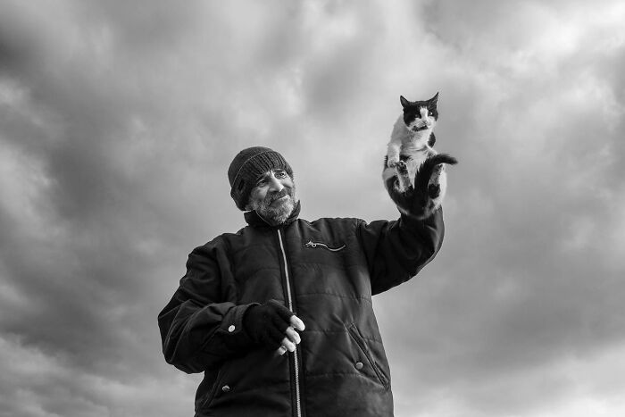 Man smiling on Istanbul streets holding a black and white cat against a cloudy sky, capturing life and cats in the city.