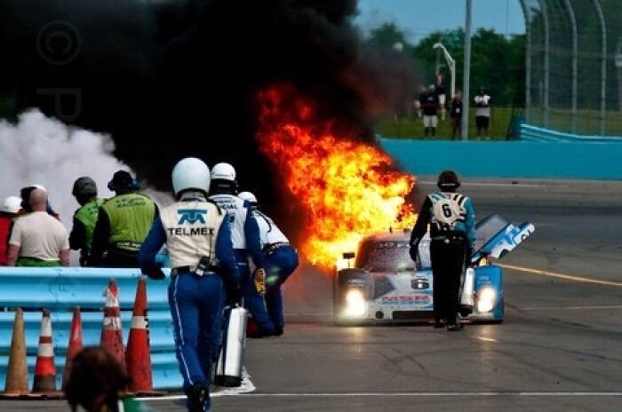 Race car engulfed in flames with crew members approaching the vehicle in a creepy and intense photo moment.