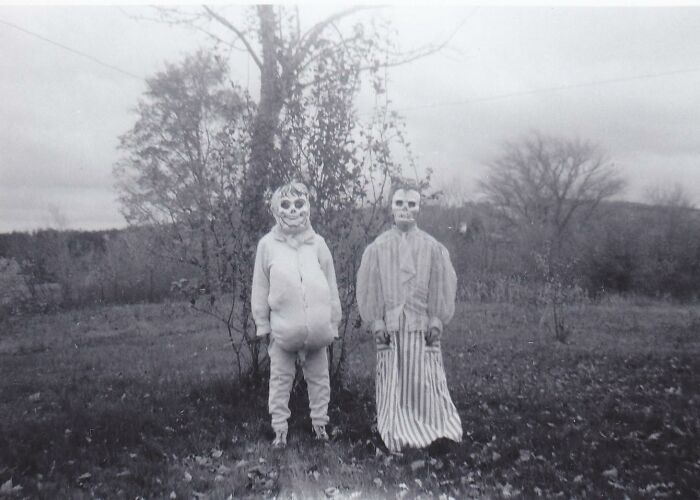Two people wearing vintage Halloween costumes with skeletal face paint standing outdoors in a spooky setting.
