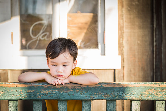 Young boy looking sad and thoughtful while leaning on a weathered green wooden railing outside a rustic building.