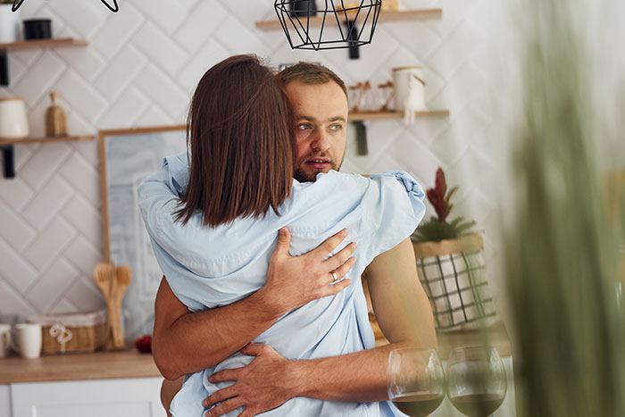 Husband and wife embracing in kitchen, husband looks upset while wife hugs him, reflecting ugly woman emotions.