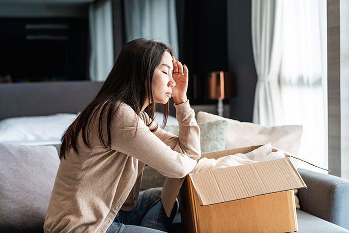 Woman sitting on couch looking stressed next to open cardboard box, reflecting a wake-up call from bumping into BF&rsquo;s grown son.