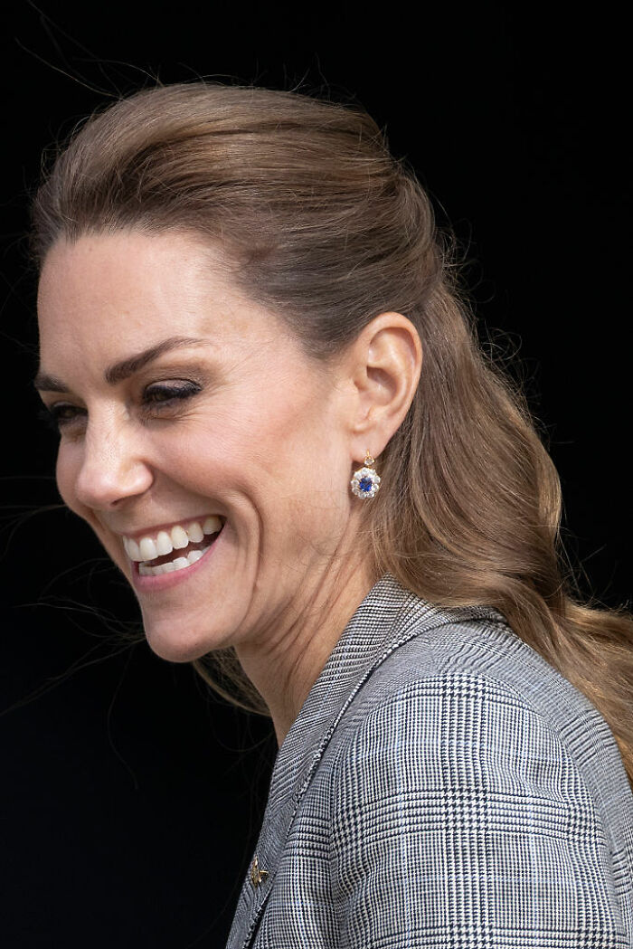 Kate Middleton smiling warmly with earrings and hair pulled back against a black background.