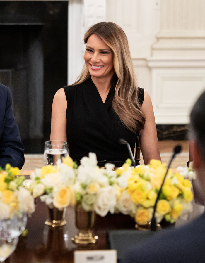 Melania Trump smiling in a black sleeveless dress, seated at a table with yellow and white floral arrangements nearby.
