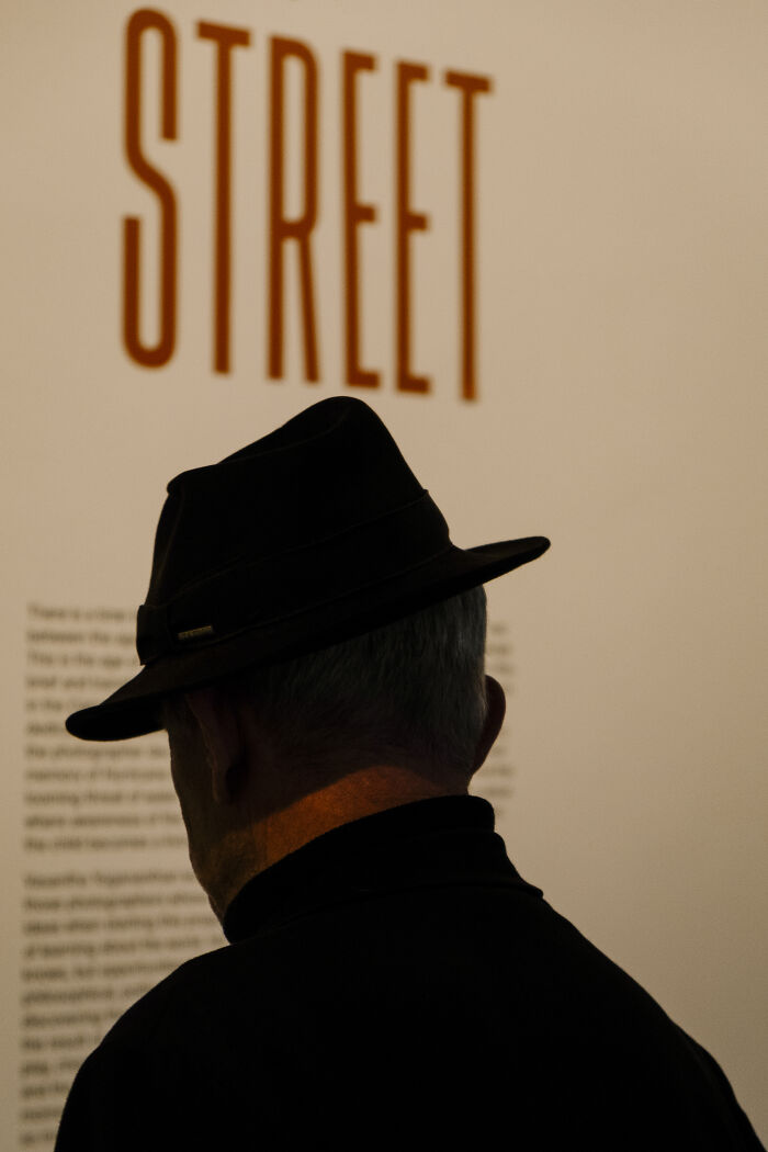 Silhouette of a man wearing a hat, standing in front of a wall with bold street photography text and blurred writing.