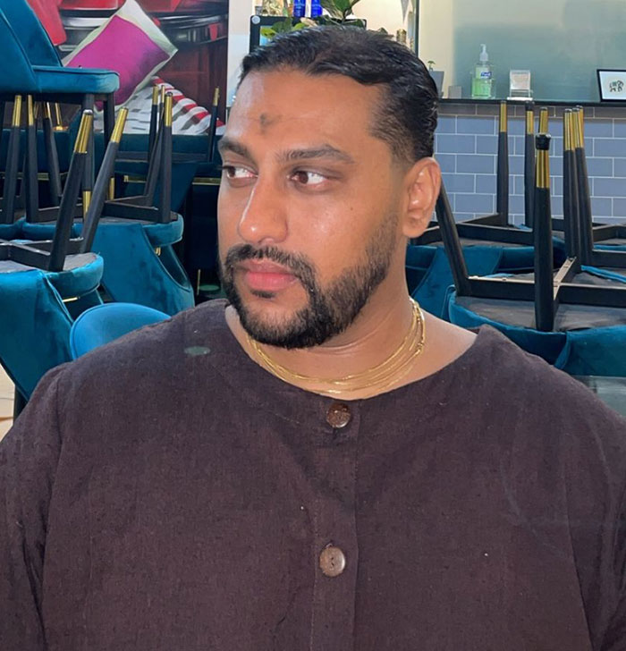 Man with beard and gold chains looking away in a room with chairs stacked upside down, related to Life Of Charlie Kirk homework outrage.