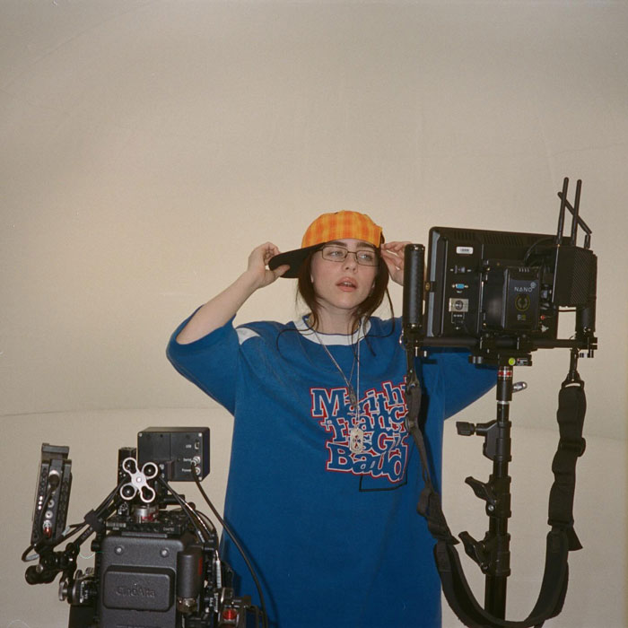 Billie Eilish on set wearing an orange cap and blue sweatshirt amid professional filming equipment and cameras.