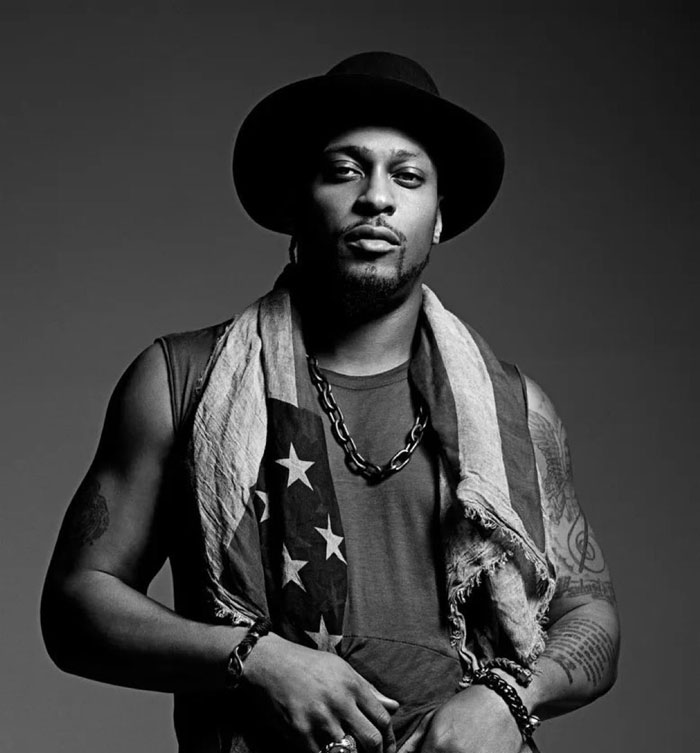 R&B legend D'Angelo wearing a hat and scarf, posing confidently with tattoos visible on his arms in black and white.