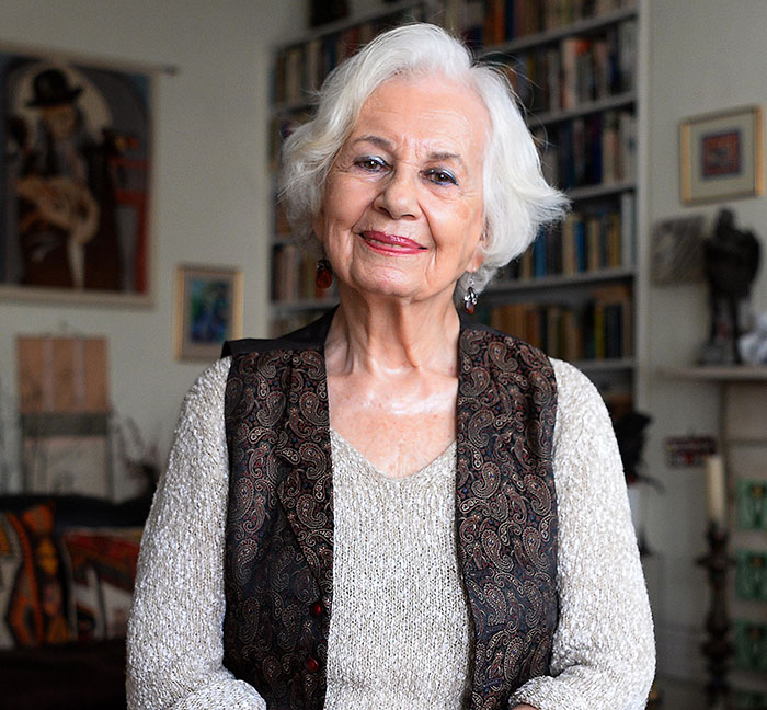 Holocaust survivor Ruth Posner smiling indoors with white hair and a patterned vest in a room filled with books.