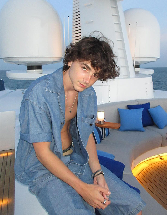 Timoth&eacute;e Chalamet sitting casually on a yacht deck near ocean, wearing denim outfit with tousled hair at dusk.