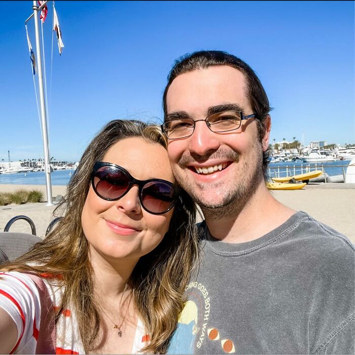 Couple smiling outdoors near a marina with boats, capturing a moment featuring pregnant Disney superfan in bright daylight.