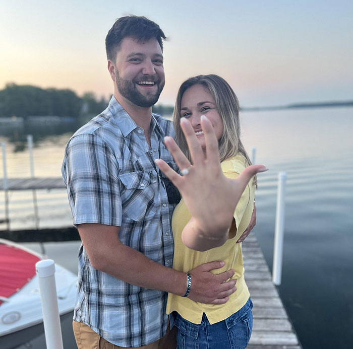Couple smiling on dock by lake, woman showing engagement ring, officials reveal cause of passing before wedding anniversary.