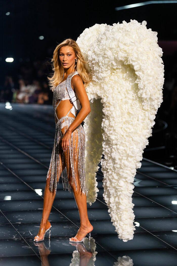 Bella Hadid walking runway wearing large white Victoria&rsquo;s Secret angel wings with silver fringe outfit on glossy catwalk.