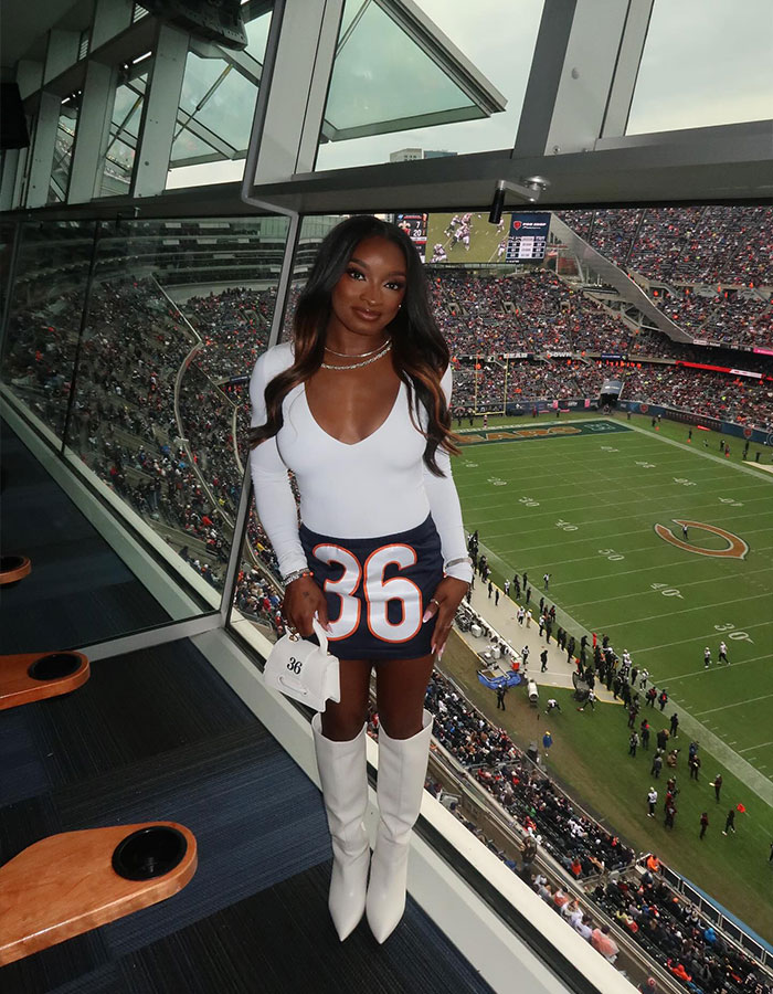 Simone Biles in a white top and boots at a football stadium wearing a skirt with number 36, fans in background.