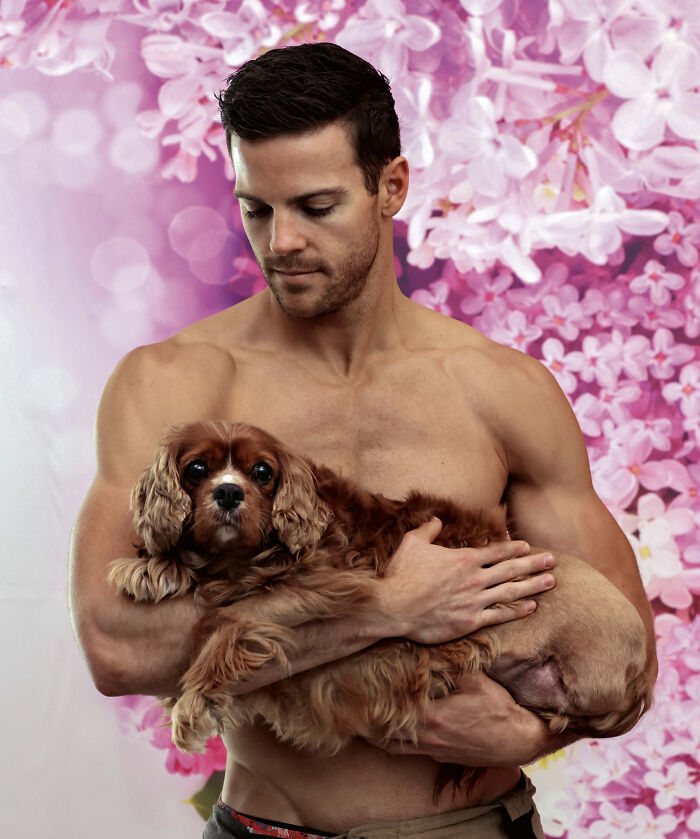 Shirtless firefighter gently holding an adorable dog with a pink floral background in an Australian calendar photoshoot.